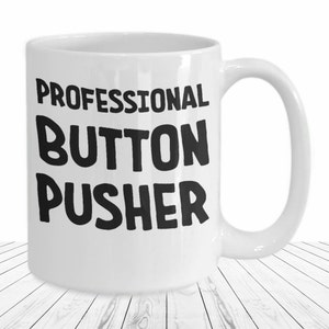 May include: White ceramic mug with a large handle. The mug features the text "PROFESSIONAL BUTTON PUSHER" in bold black letters. The mug is sitting on a white wooden surface.