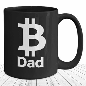 Könnte beinhalten: Schwarze Keramik-Kaffeetasse mit einem weißen Bitcoin-Symbol und dem Wort "Dad" darauf gedruckt.