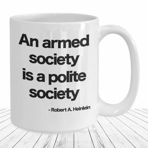 May include: White ceramic coffee mug with black text that reads "An armed society is a polite society - Robert A. Heinlein".