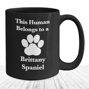May include: Black coffee mug with white text that reads "This Human Belongs to a Brittany Spaniel" and a white paw print.