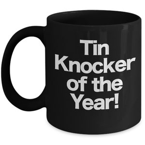 May include: Black ceramic coffee mug with white text that reads "Tin Knocker of the Year!"