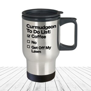 May include: A stainless steel travel mug with a black lid and handle. The mug has a white background with black text that reads "Curmudgeon To Do List: Coffee No Get Off My Lawn".