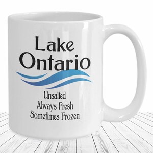 May include: White ceramic coffee mug with a blue and white wave design and the text "Lake Ontario Unsalted Always Fresh Sometimes Frozen".