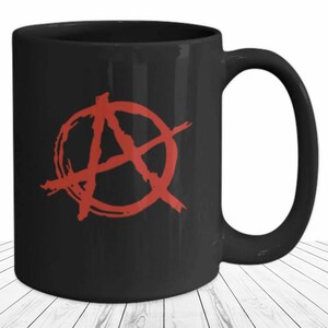 May include: Black ceramic mug featuring a red anarchy symbol. The symbol is a stylized letter "A" inside a circle with radiating lines. The mug has a curved handle and a glossy finish.