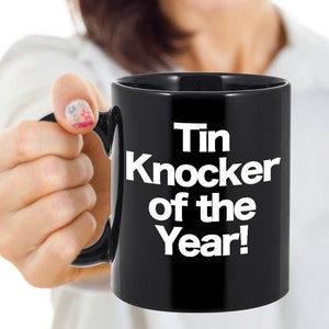 May include: Black coffee mug with white text that reads "Tin Knocker of the Year!"