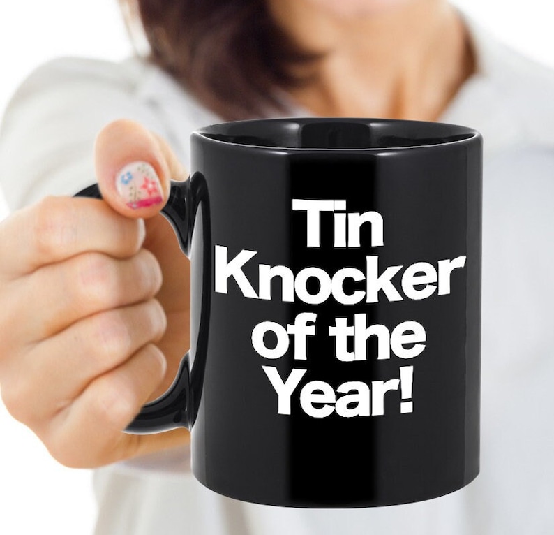 May include: Black coffee mug with white text that reads "Tin Knocker of the Year!"