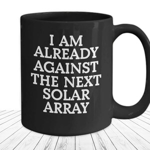 May include: Black ceramic coffee mug with white text that reads "I am already against the next solar array".