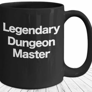 Könnte beinhalten: Schwarze Keramik-Tasse mit großem Henkel. Auf der Tasse steht in weißer Schrift "Legendary Dungeon Master" in einer fetten, serifenlosen Schrift. Die Tasse steht auf einer weißen Holzoberfläche.