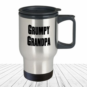 May include: A stainless steel travel mug with a black lid and handle. The words "GRUMPY GRANDPA" are printed in bold black letters on the front of the mug. The mug is designed to keep beverages hot or cold.