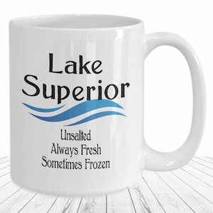 May include: A white ceramic mug featuring the black text Lake Superior above a blue wave graphic. Below the wave, the black text reads Unsalted, Always Fresh, Sometimes Frozen. The mug has a standard handle and a smooth, glossy finish.