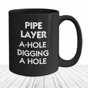 May include: Black coffee mug with white text that reads "PIPE LAYER A-HOLE DIGGING A HOLE".