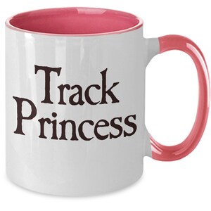May include: White ceramic coffee mug with a pink rim and handle. The mug has black text that reads "Track Princess".