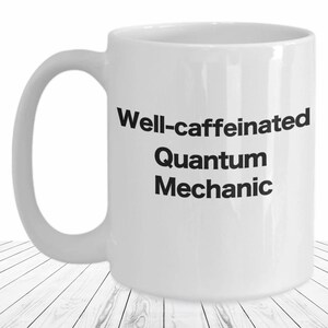 May include: White ceramic coffee mug with black text that reads "Well-caffeinated Quantum Mechanic".