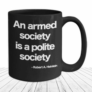 May include: Black coffee mug with white text that reads "An armed society is a polite society - Robert A. Heinlein"