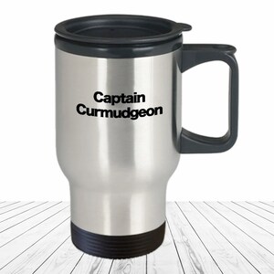 May include: A stainless steel travel mug with a black lid and handle. The mug has the text "Captain Curmudgeon" printed on the front.