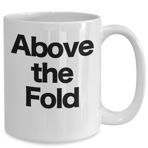May include: White ceramic coffee mug with black text that reads "Above the Fold".