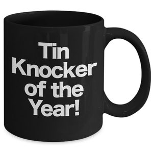 May include: Black ceramic coffee mug with white text that reads "Tin Knocker of the Year!"