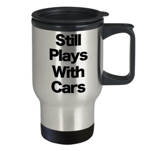 Könnte beinhalten: Eine silberne Reise-Thermoskanne mit schwarzem Deckel und Griff. Auf der Kanne steht der Text "Still Plays With Cars".