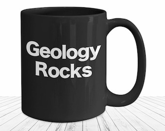 May include: A black ceramic mug featuring the white text Geology Rocks printed on the front. The mug has a standard cylindrical shape with a curved handle on the right side, providing a simple and functional design for holding hot or cold beverages.