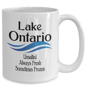 May include: White ceramic coffee mug with a black and blue design. The mug features the text "Lake Ontario" with a blue wave design and the text "Unsalted Always Fresh Sometimes Frozen".