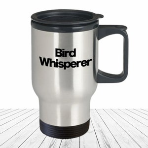 May include: A stainless steel travel mug with a black lid and handle. The mug has the text "Bird Whisperer" printed on the front in black.