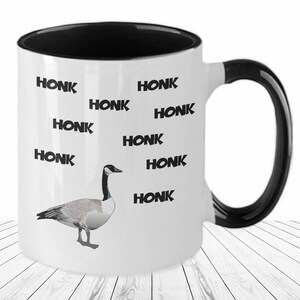 May include: White ceramic coffee mug with a black rim and handle. The mug features a black and white illustration of a goose and the word "HONK" repeated multiple times.