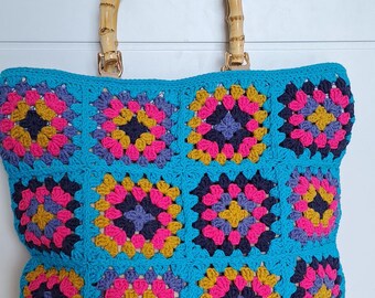 May include: A crochet tote bag featuring a vibrant granny square pattern in shades of turquoise, hot pink, mustard yellow, and dark navy blue. The bag is constructed with a turquoise border and includes two rounded bamboo handles.