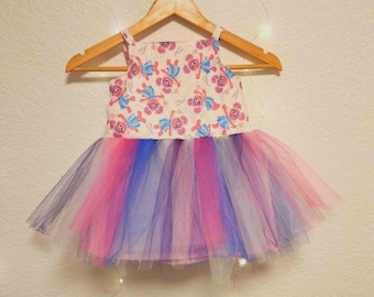 May include: A white tulle tutu dress with a colorful rainbow tulle skirt. The bodice of the dress has a white background with a pattern of pink and blue cartoon characters.