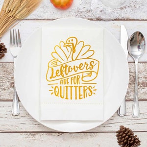 Peut inclure: Une serviette blanche avec un motif de dinde dor&eacute; et le texte "Leftovers are for Quitters" est pos&eacute;e sur une assiette blanche. Une fourchette, un couteau et une cuill&egrave;re sont dispos&eacute;s. D&eacute;cor de table avec pomme de pin et &eacute;pis de bl&eacute;.