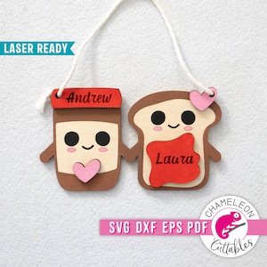 May include: Wooden laser-cut ornaments depicting a jar of spread and a slice of toast, each with smiling faces and holding hands. The jar has the name "Andrew" and the toast has "Laura". Both have pink hearts. A white string is attached for hanging.