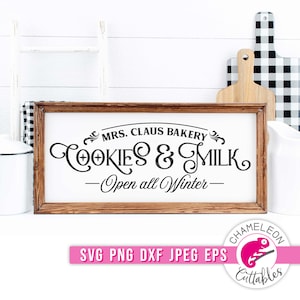 Op de afbeelding: Een rechthoekig houten bord met de tekst "MRS. CLAUS BAKERY COOKIES & MILK - Open all Winter-" in zwart schrift. Het bord is omlijst in donkerbruin hout. Een witte achtergrond met een ladder, snijplanken en een kan is zichtbaar.
