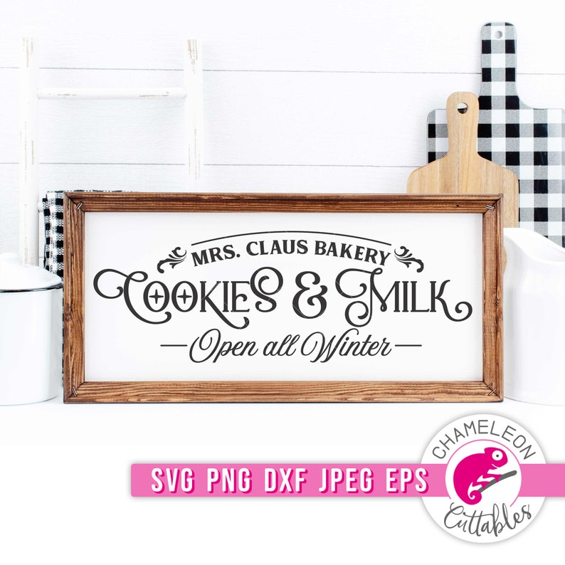 Op de afbeelding: Een rechthoekig houten bord met de tekst "MRS. CLAUS BAKERY COOKIES & MILK - Open all Winter-" in zwart schrift. Het bord is omlijst in donkerbruin hout. Een witte achtergrond met een ladder, snijplanken en een kan is zichtbaar.