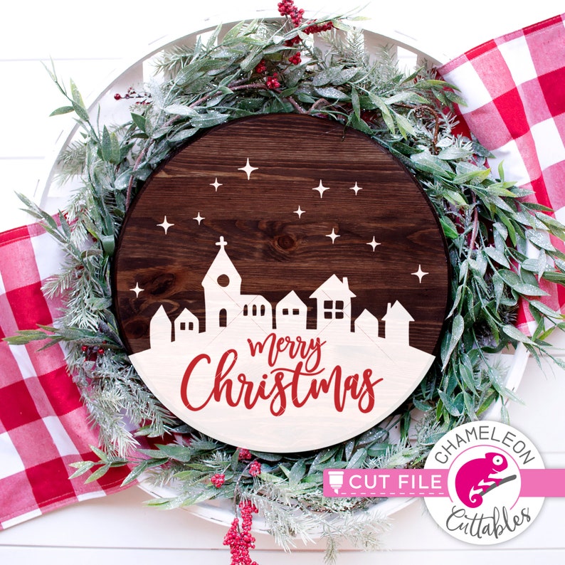May include: A round wooden sign with a white silhouette of a town with a church and the words "Merry Christmas" in red lettering. The sign is decorated with white stars and is surrounded by a green wreath with red berries.
