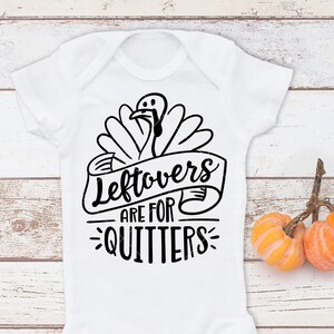 Peut inclure: Body pour b&eacute;b&eacute; blanc avec un dessin de dinde noire et l'inscription "Leftovers are for Quitters". Deux petites citrouilles oranges en arri&egrave;re-plan. Un design festif pour les f&ecirc;tes de Thanksgiving.