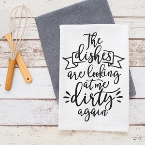 May include: White kitchen towel with the text "The dishes are looking at me dirty again" in black script. A gray dish towel is underneath. A whisk and spatula with wooden handles are on the left.