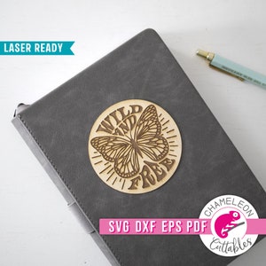 K&ouml;nnte beinhalten: Ein Holzrund mit einem Schmetterlingsdesign und den Worten "WILD AND FREE" eingraviert. Das Holzrund befindet sich auf einem grauen Notizbuch. Das Bild enth&auml;lt auch einen Stift und den Text "LASER READY".