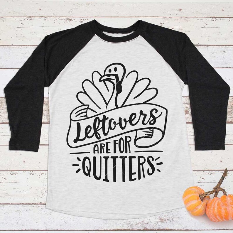 Peut inclure: Un t-shirt de style raglan avec un corps blanc et des manches gris fonc&eacute;. Le t-shirt pr&eacute;sente un dessin noir d'une dinde avec le texte "Leftovers are for Quitters" sur une banni&egrave;re. Deux petites citrouilles sont dans le coin inf&eacute;rieur droit.