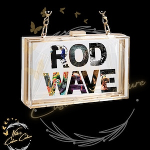 May include: A clear acrylic clutch with gold-tone hardware and a chain strap. The front of the clutch features the words "ROD WAVE" with album art images inside the letters. The clutch is rectangular and has a clasp closure.