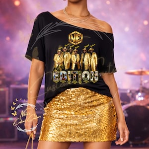 May include: Black off-the-shoulder t-shirt featuring a gold graphic of the group New Edition. The graphic includes the band's logo and members' names. The model is wearing a gold sequin mini skirt. The shirt is from Nikki's Custom Couture.