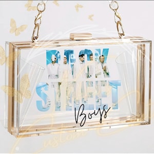 Puede incluir: Bolso de mano transparente de acrílico con detalles dorados y una correa de cadena. El bolso presenta un gráfico de los Backstreet Boys con su nombre en un diseño azul y blanco. La palabra "Boys" está escrita en cursiva.