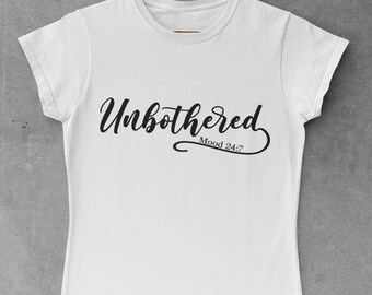 Peut inclure: T-shirt blanc avec le mot "Unbothered" en écriture noire. En dessous, l'expression "Mood 24:7" est imprimée dans une police plus petite. Le t-shirt est suspendu à un cintre cuivré sur un fond gris.