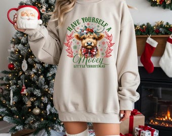 May include: Beige sweatshirt with a graphic of a cow wearing a plaid bow tie and a floral crown. The text reads "Have Yourself a Mooey Little Christmas." The sweatshirt is part of a Christmas-themed collection.