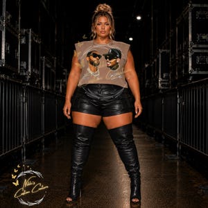 May include: A woman wearing a sleeveless brown t-shirt with a graphic of two men, black leather shorts, and black thigh-high boots. The boots have open toes and high heels. The background is dark with stage equipment.