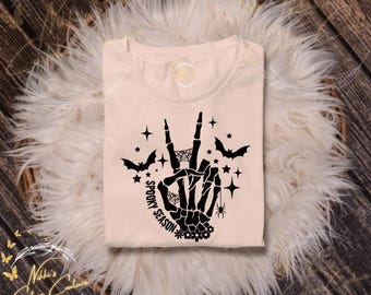 May include: A light beige t-shirt featuring a black skeleton hand making a peace sign, with the words "Spooky Season" and bat and star graphics. The shirt is displayed on a fluffy white surface.