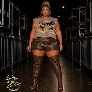 May include: A woman wearing a brown sleeveless top with a graphic of two men, brown leather shorts, and brown thigh-high boots. The top has a crew neck and the shorts have pockets. The boots have a stiletto heel.