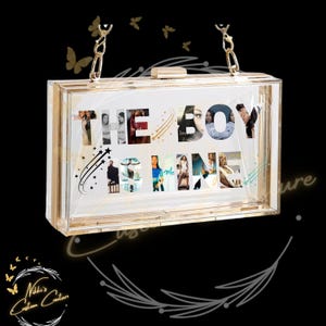 May include: A clear acrylic clutch with gold-tone hardware and a chain strap. The front of the clutch features the words "THE BOY IS MINE" with images of people inside the letters. Decorative butterflies are in the background.