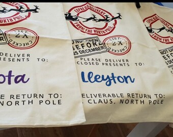 May include: Off-white Christmas gift sacks with red and black stamped designs, including Santa's sleigh and reindeer. Each sack is personalized with a name in a different color: Dakota, Lleyton, and Shelby.