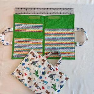 May include: A fabric organizer with a green interior featuring multi-colored wavy striped pockets and a white exterior patterned with cartoon characters, rockets, and planets. A clear ruler shows the organizer measures 17 inches in width.