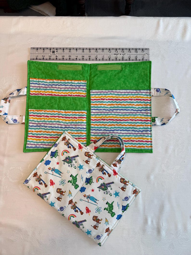 May include: A fabric organizer with a green interior featuring multi-colored wavy striped pockets and a white exterior patterned with cartoon characters, rockets, and planets. A clear ruler shows the organizer measures 17 inches in width.