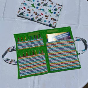 May include: A fabric carrying case with a white exterior featuring a colorful cartoon character print and a bright green interior. The case unfolds to reveal multiple slots holding colored pencils and a pocket containing a limited edition coloring book.